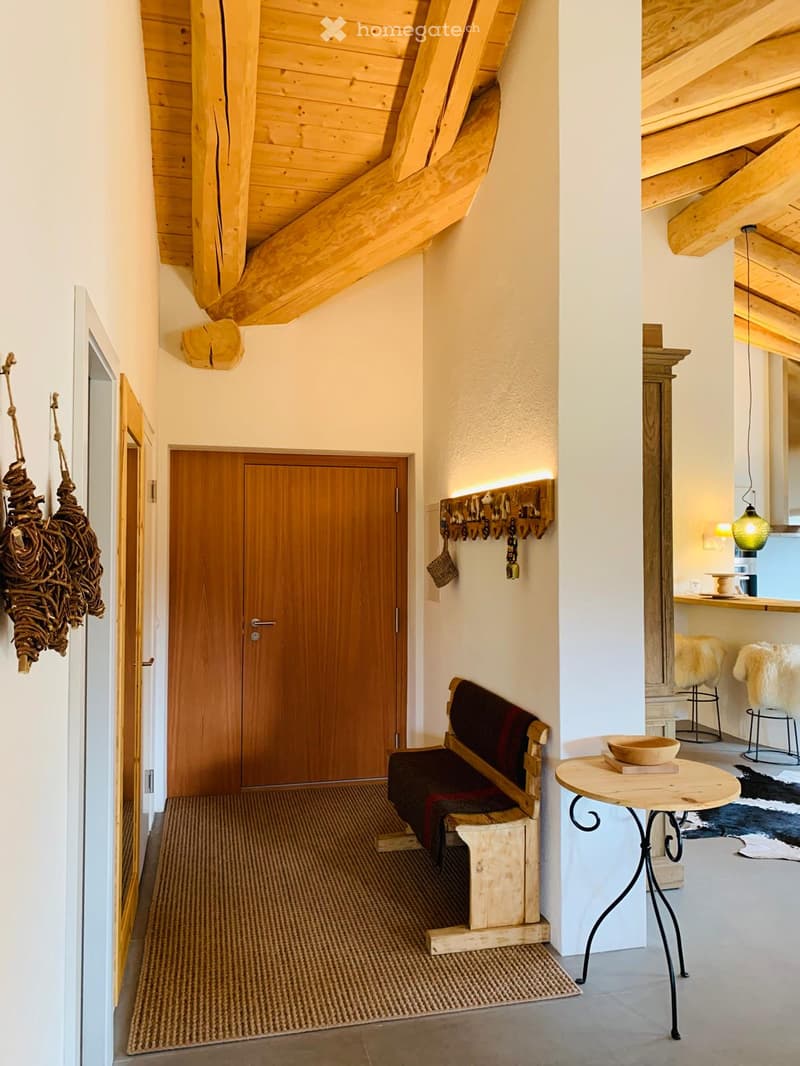 Cozy hallway with wooden ceiling beams, a wooden bench, a small round table, and decorative items including a hanging rope and fur-covered chairs visible in the adjacent room.