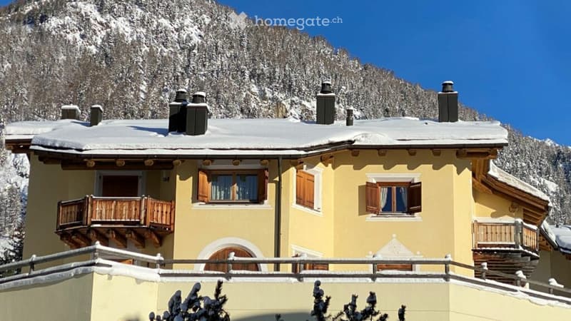 Yellow two-story house with wooden balconies and shutters, set against a snowy mountain backdrop.