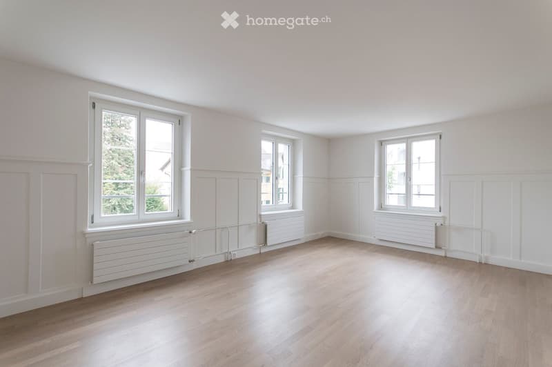 Empty room with light wooden flooring, white walls, and multiple windows with white frames. Radiators are visible beneath the windows.