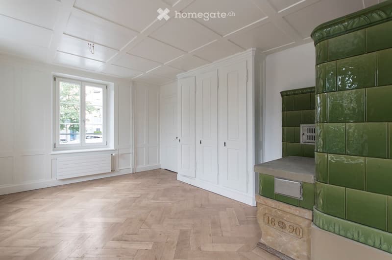 Empty room with wooden flooring, white walls, large window, and a green tiled fireplace.
