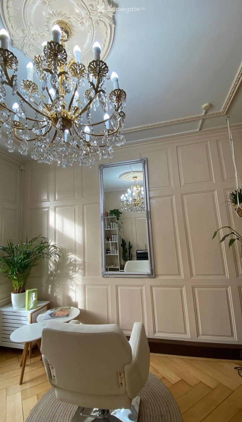 Elegant room with a large ornate chandelier, a silver-framed mirror, a white chair, a small round table, and a potted plant on a white cabinet.