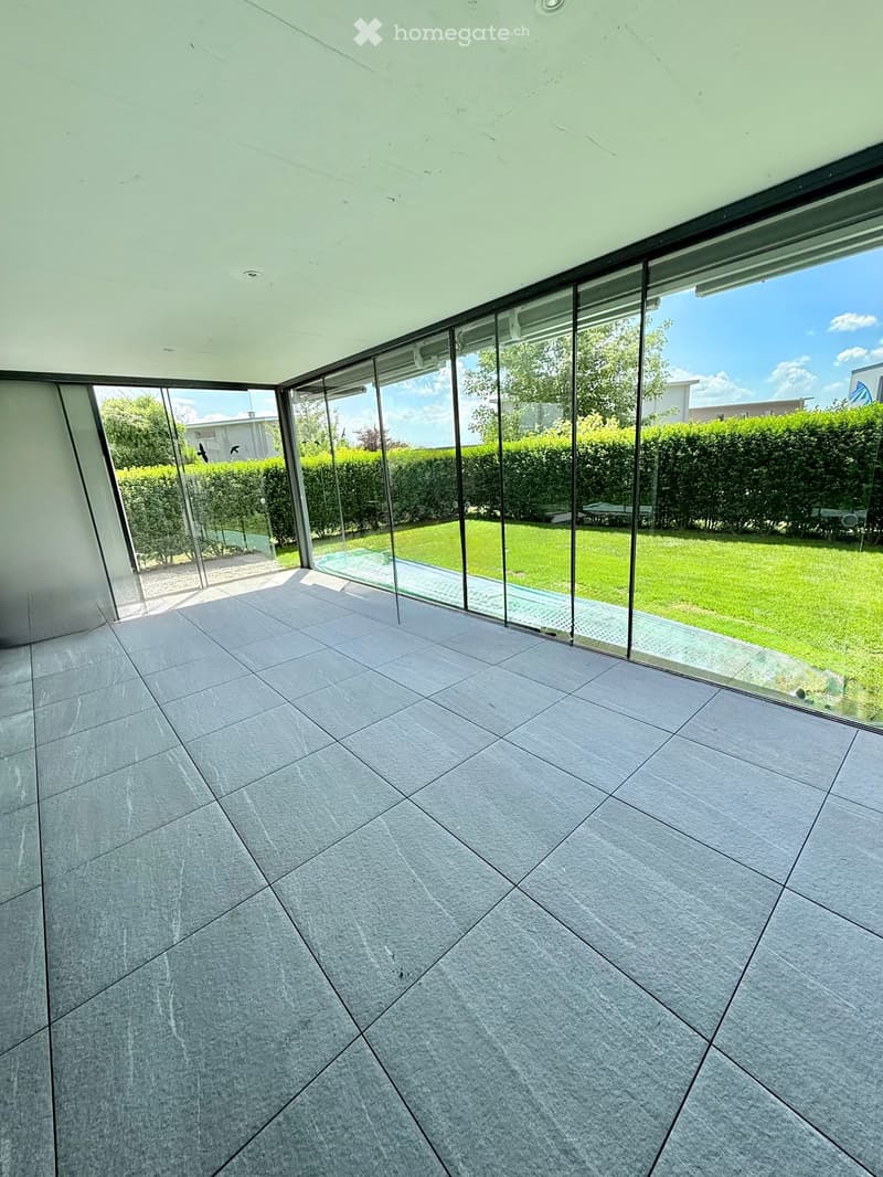 Empty room with large glass walls and a view of a green garden, featuring a tiled floor and modern ceiling lighting.