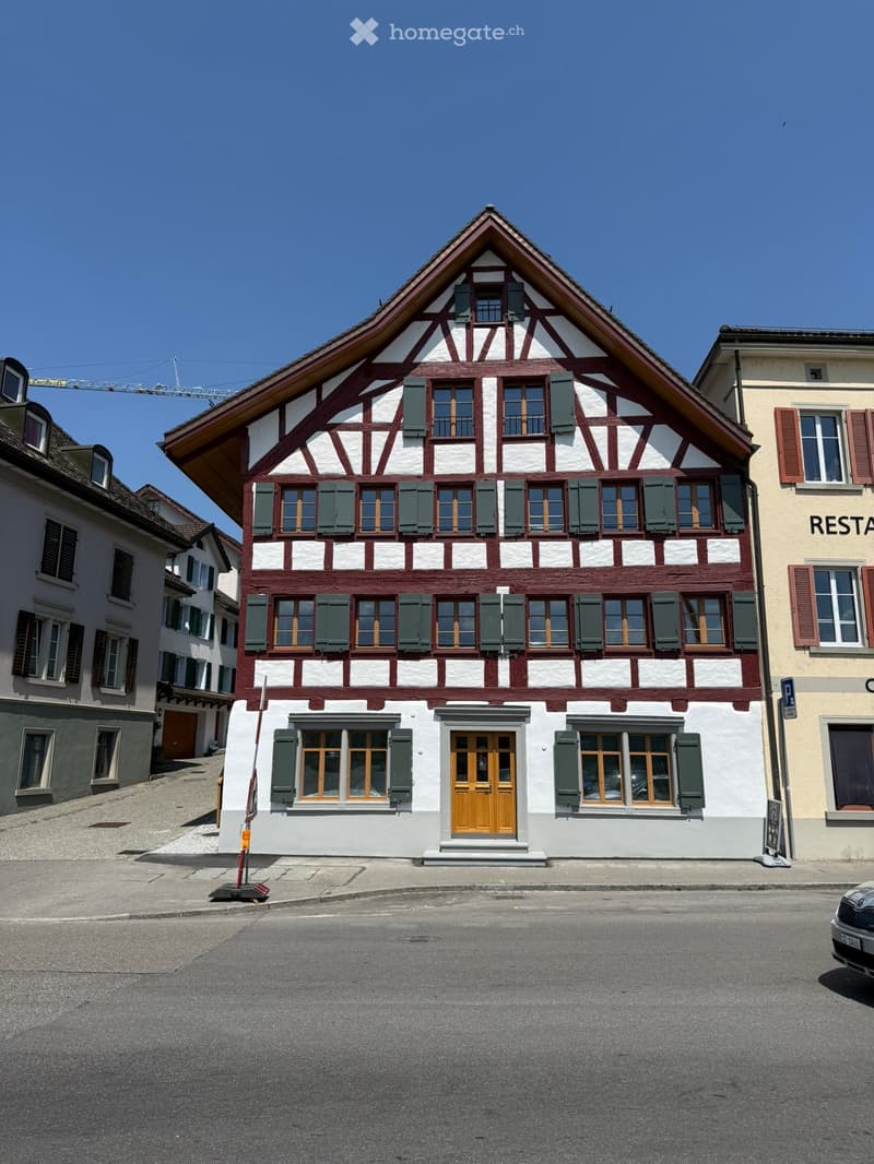 Traditional Swiss half-timbered building with white and red facade, green shutters, and a steeply pitched roof.