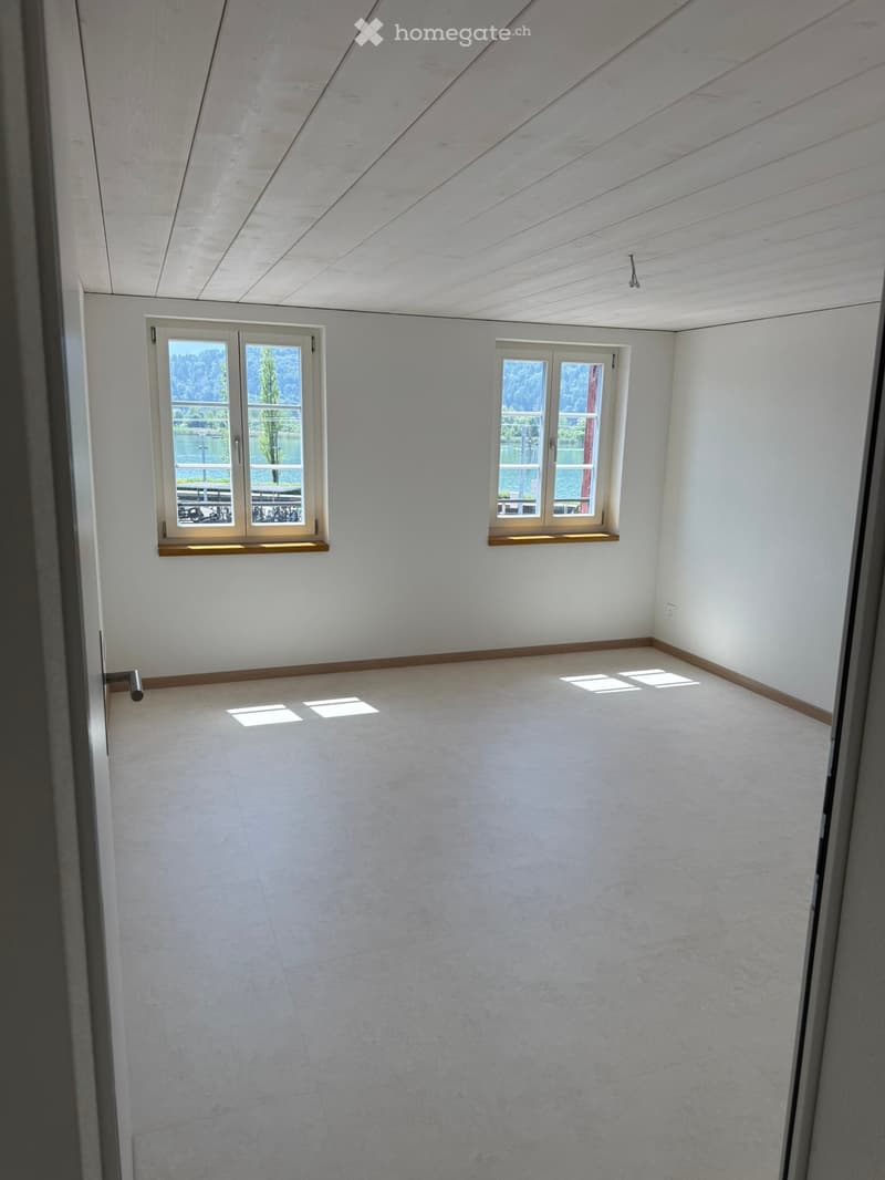 Empty room with white walls, light-colored tiled floor, and two windows offering a scenic view of a lake and trees.