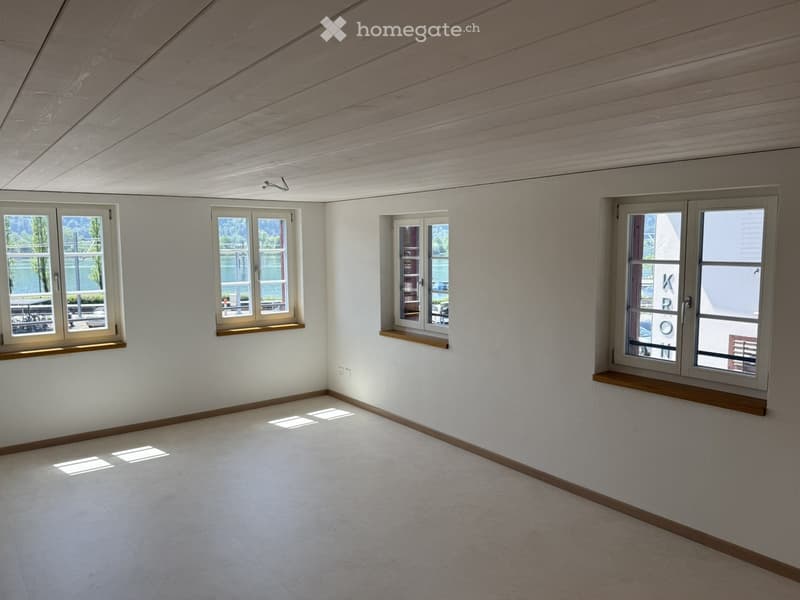 Empty room with white walls, light wooden ceiling, and multiple windows with wooden sills. The floor is covered with light-colored carpeting.