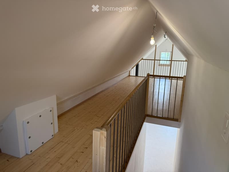 Attic corridor with wooden flooring, white walls, and a metal railing leading to a windowed area.