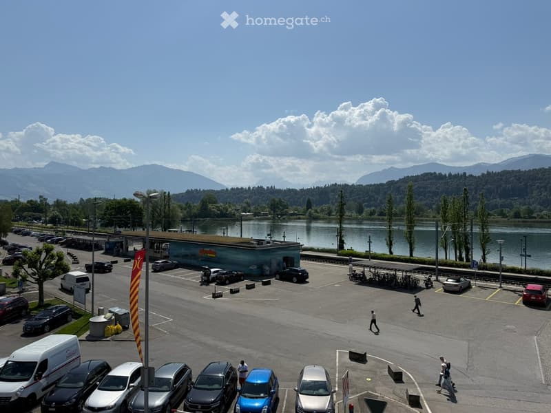 Parking lot with several cars, a small building labeled "migrolino", and a body of water with mountains in the background.