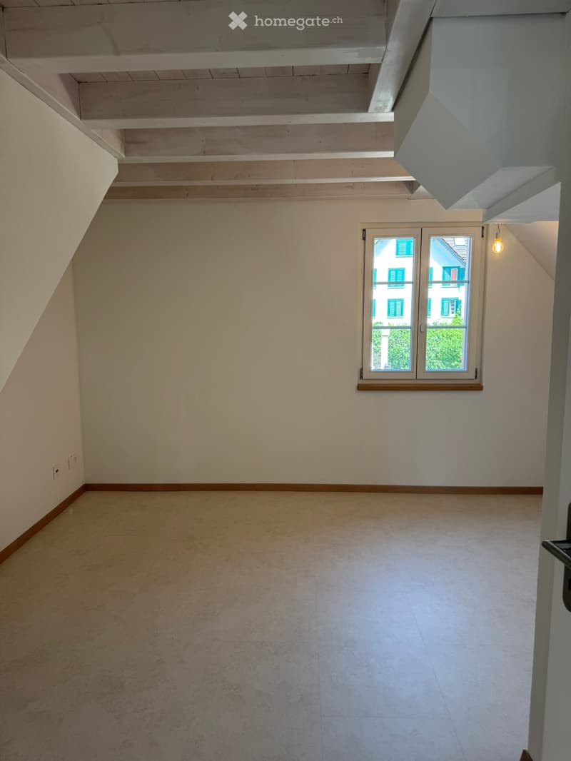 Empty attic room with wooden ceiling beams, light-colored walls, and a single window showing a view of neighboring buildings.
