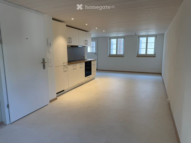 Empty room with light-colored walls, carpeted floor, and a small kitchen area featuring white cabinets, a countertop, and an oven. Two windows provide natural light.