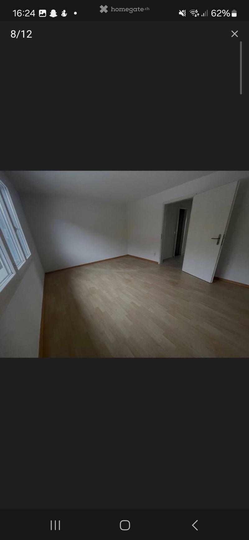 Empty room with wooden flooring, white walls, and a large window on one side. The room has a door leading to another space.