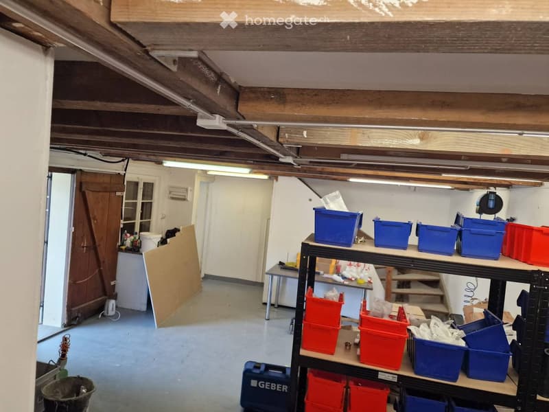 A spacious, unfinished garage with exposed wooden beams and a concrete floor. Shelves hold various boxes and tools, and a small table and stool are visible.