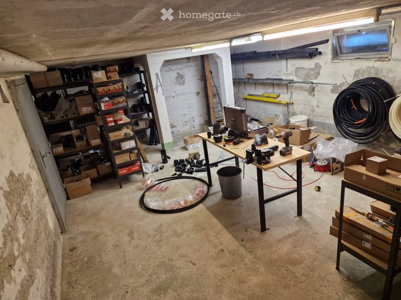Workshop or storage area with concrete floor, exposed wooden beams, shelves filled with tools and supplies, work tables with tools, and various equipment.