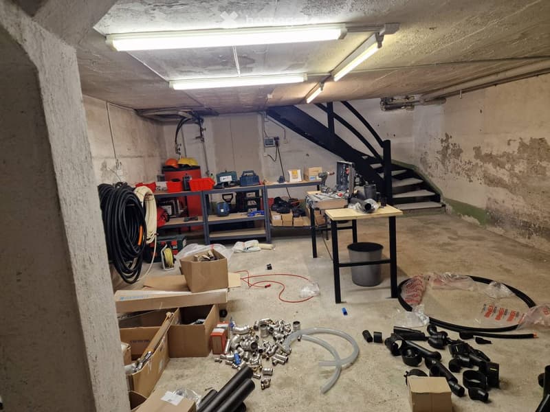 A cluttered basement or utility room with shelves holding tools and supplies, a workbench, and various pipes and cables on the floor.