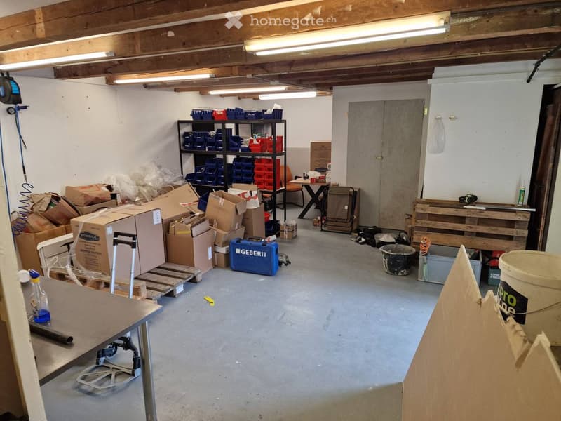 Empty storage room with exposed wooden beams, fluorescent lighting, shelves, and various storage items including boxes, tools, and containers.