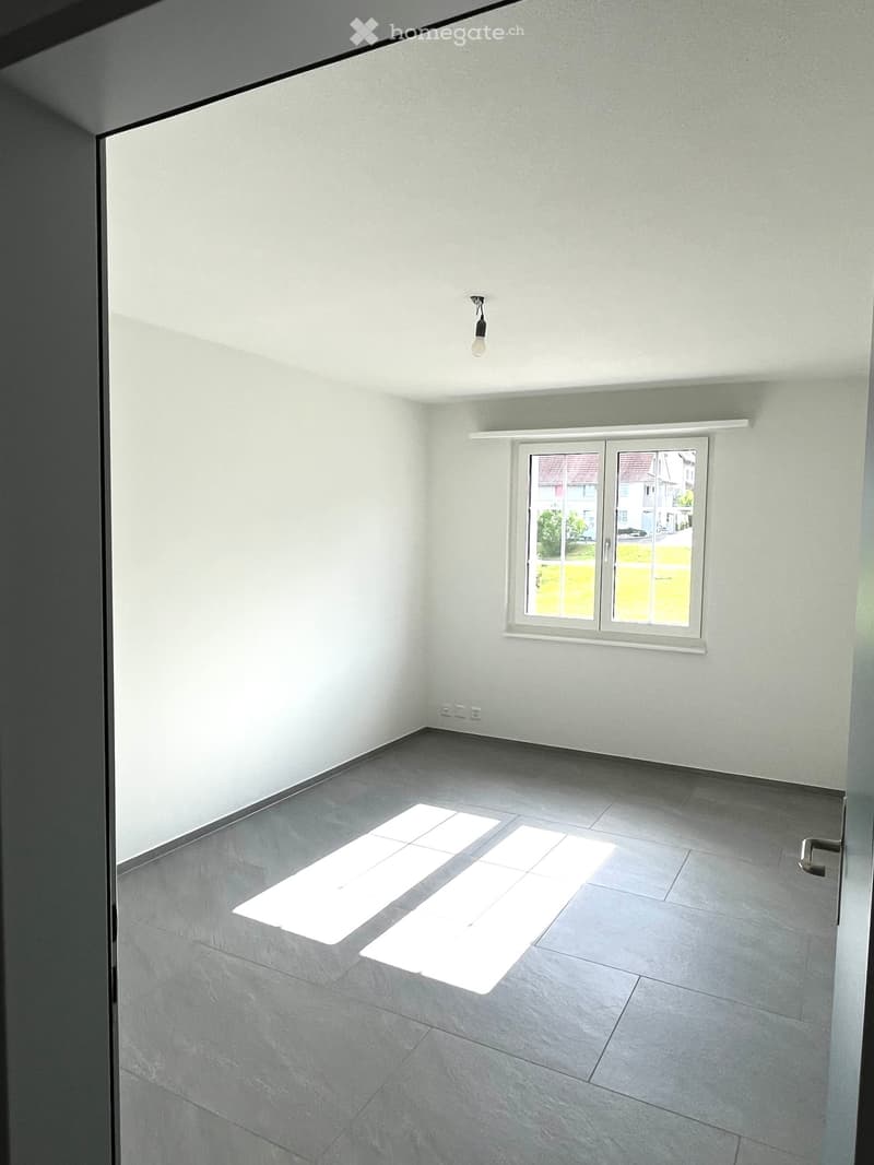 Empty room with grey tiled floor, white walls, and a single window showing a view of the street outside. The room has a modern, minimalist design with a visible light fixture on the ceiling.