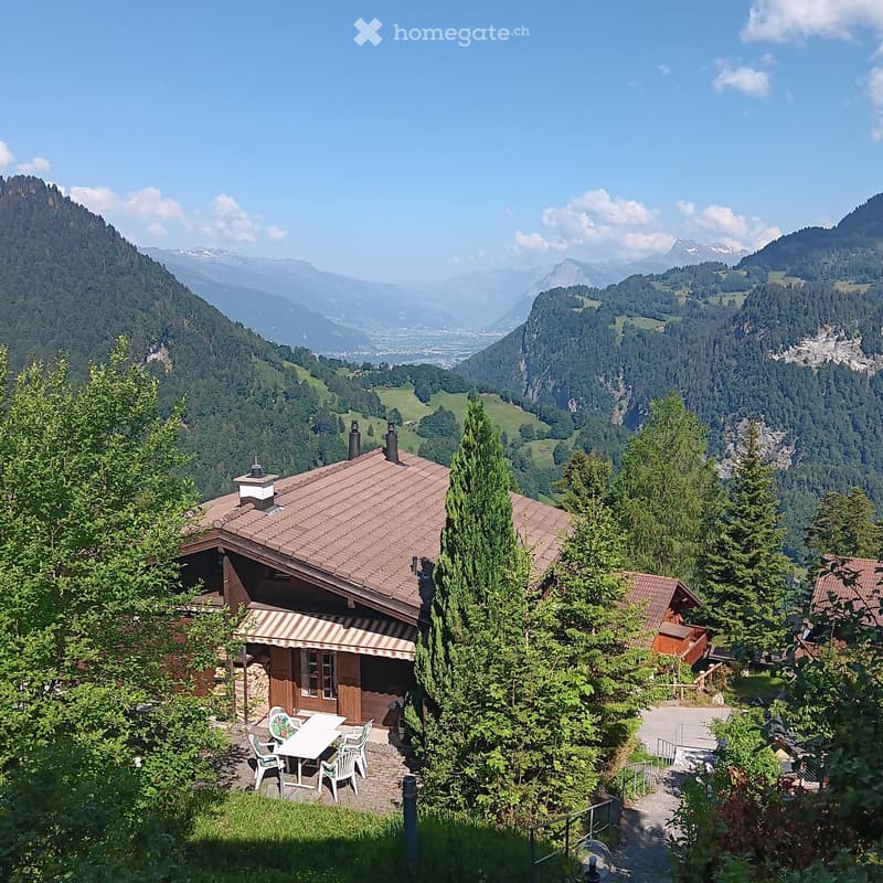 A rustic wooden house with a tiled roof, surrounded by lush greenery and offering a panoramic mountain view.