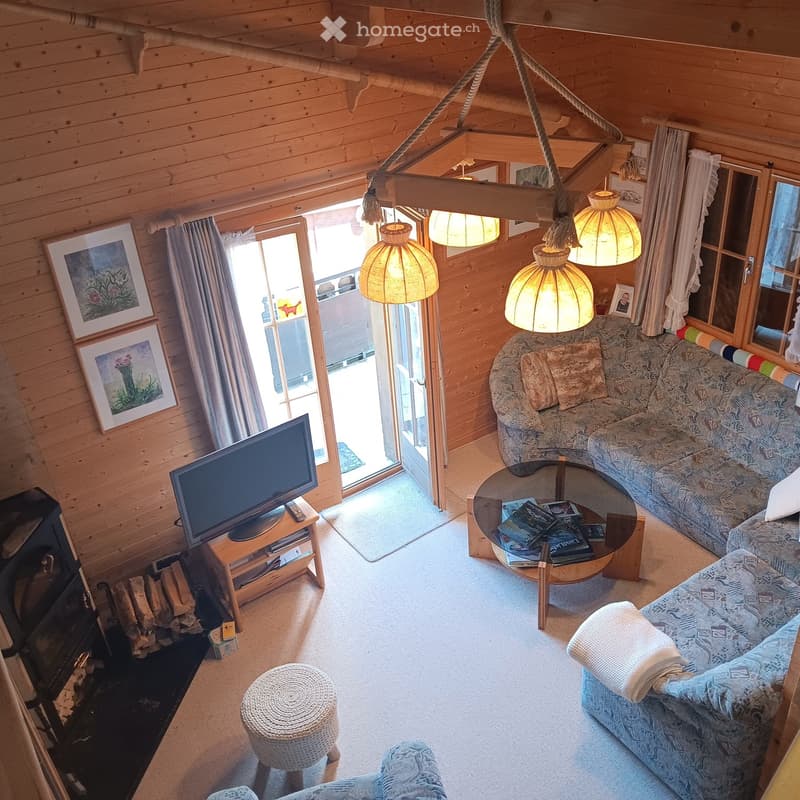 Cozy living room with wooden walls, exposed beams, and warm lighting. Features a patterned sofa, coffee table with books, a round wooden table, a television on a stand, and a woven stool.