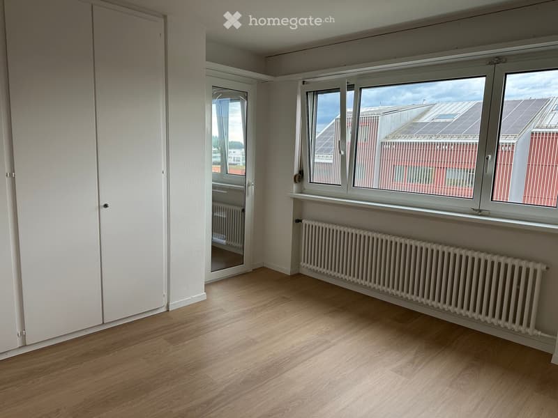 Empty room with light wooden flooring, white walls, large windows, radiator, and a mirrored door leading to another space.