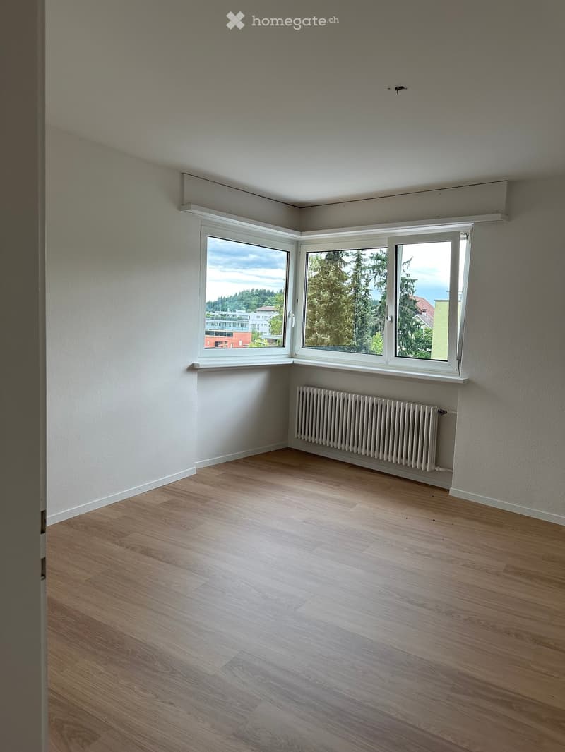 Empty room with wooden flooring, white walls, large windows, and a radiator under the windows.