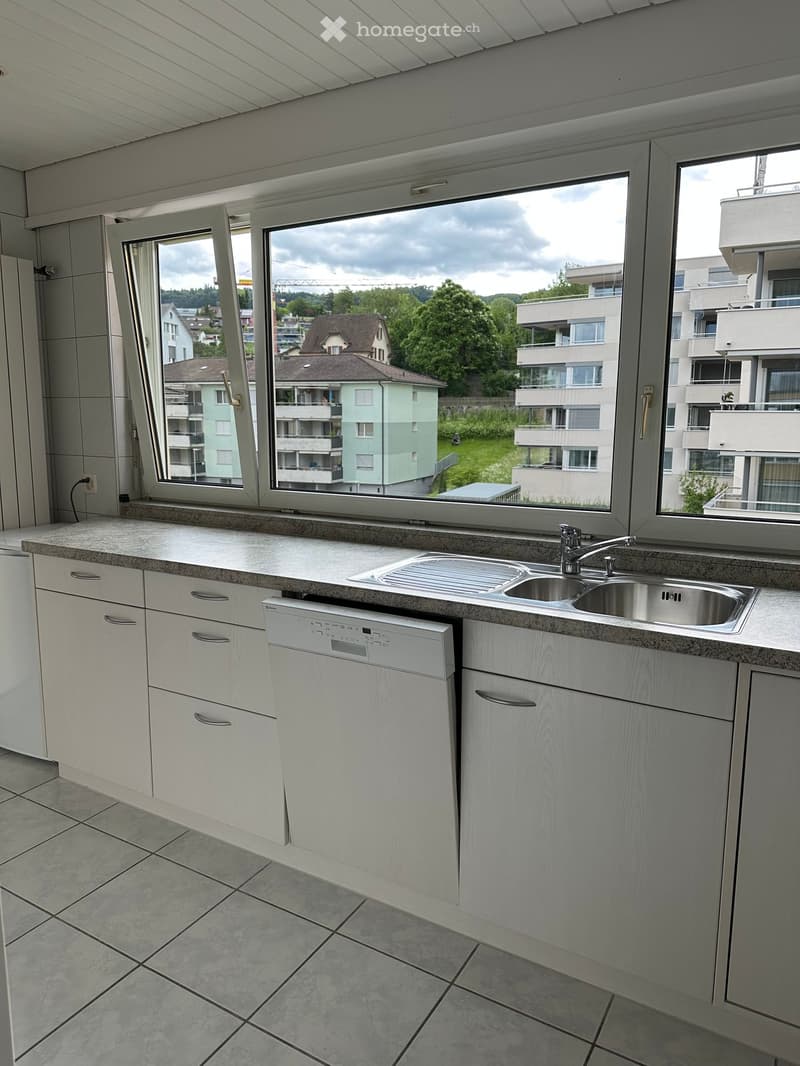 Modern kitchen with white cabinets, granite countertop, stainless steel sink, and built-in dishwasher. Large windows offer a view of the surrounding area.