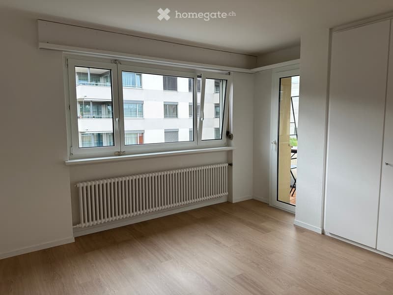 Empty room with wooden flooring, white walls, large window with radiator below, and an open doorway leading to another space.