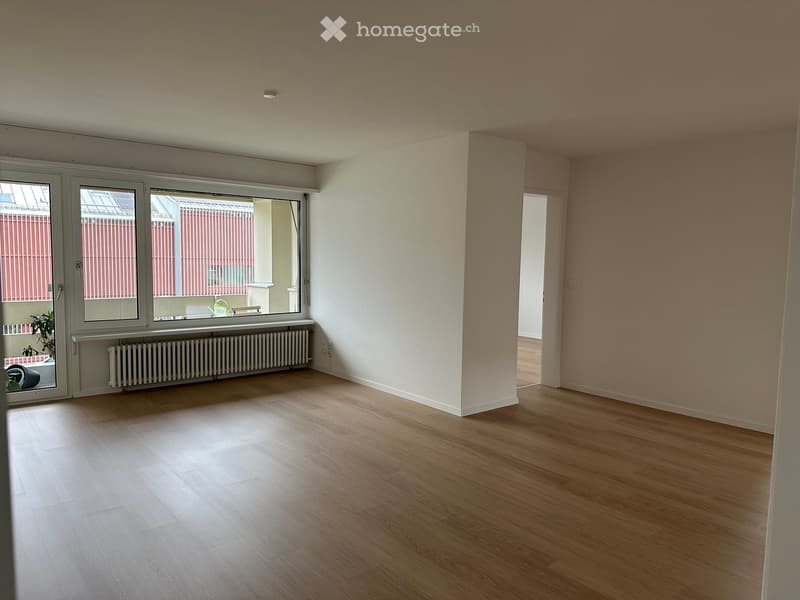 Empty room with wooden flooring, large windows, radiator, and a door leading to another space.