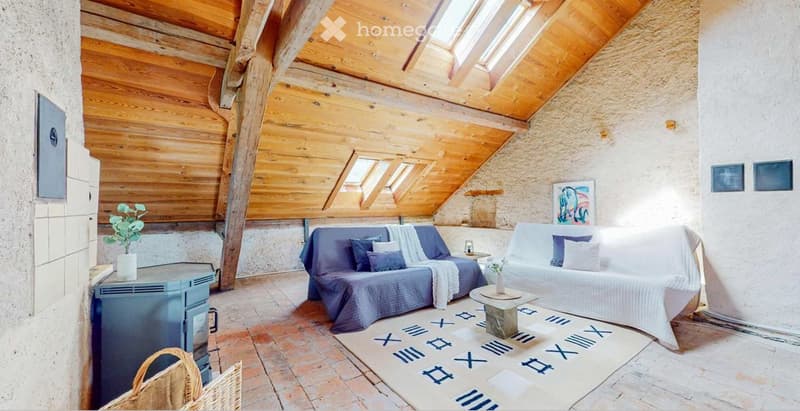 Cozy attic living space with wooden beams, skylights, exposed stone walls, and a fireplace. Features a blue sofa, white sofa, coffee table, side table, and decorative vases.