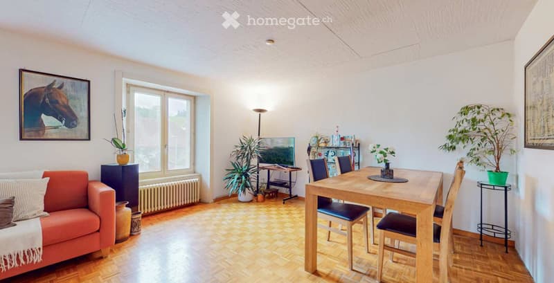 Bright living room with wooden flooring, large window, orange sofa, wooden dining table with chairs, potted plants, and a television on a stand.