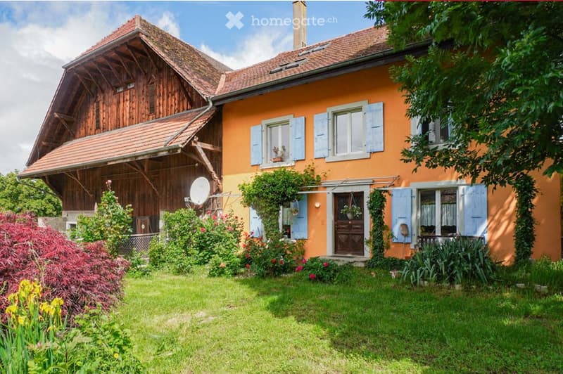 A two-story house with a wooden extension, orange facade, and multiple windows with shutters. The house has a well-maintained garden with various plants and flowers.
