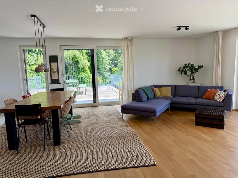 Modern living and dining area with a grey sectional sofa, wooden coffee table, dining table with chairs, and large windows leading to a balcony.