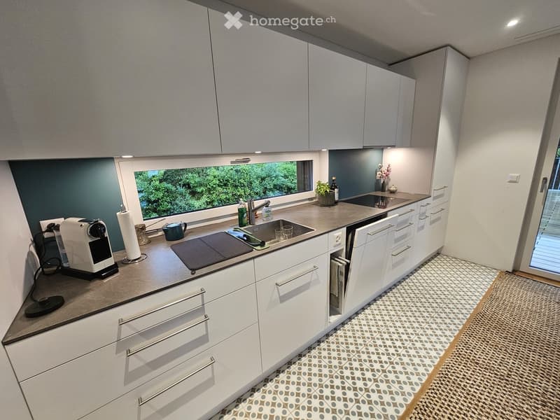 Modern kitchen with white cabinets, stainless steel appliances, and a large window overlooking greenery. Features include a sink, coffee maker, oven, and various kitchen items on the counter.