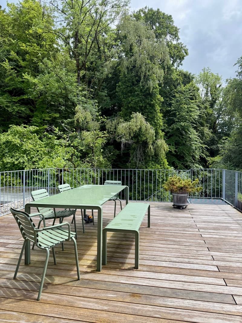 Wooden deck with green metal furniture, including a table, chairs, and a bench, surrounded by a metal railing and lush greenery.