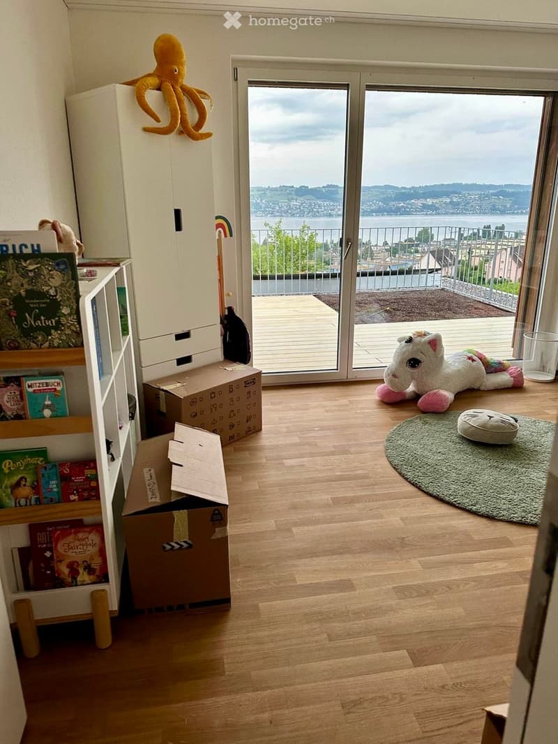 A room with wooden flooring, a large window offering a scenic view, a bookshelf filled with books, and several toys including a plush octopus and unicorn, along with a small rug and cat bed.