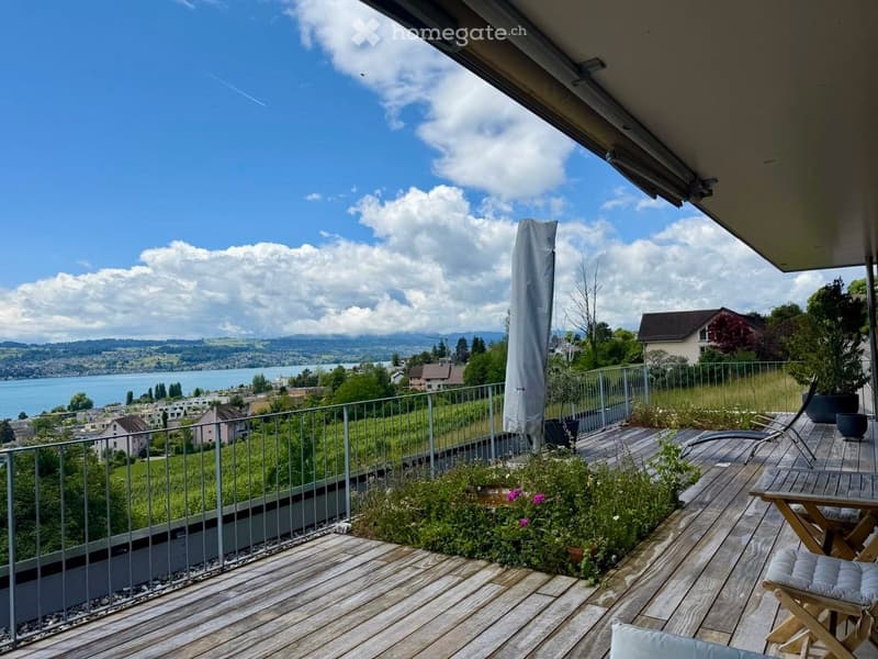 Balcony with wooden decking, metal railing, potted plants, lounge chair, small table, and umbrella cover, overlooking a scenic lake and mountains.