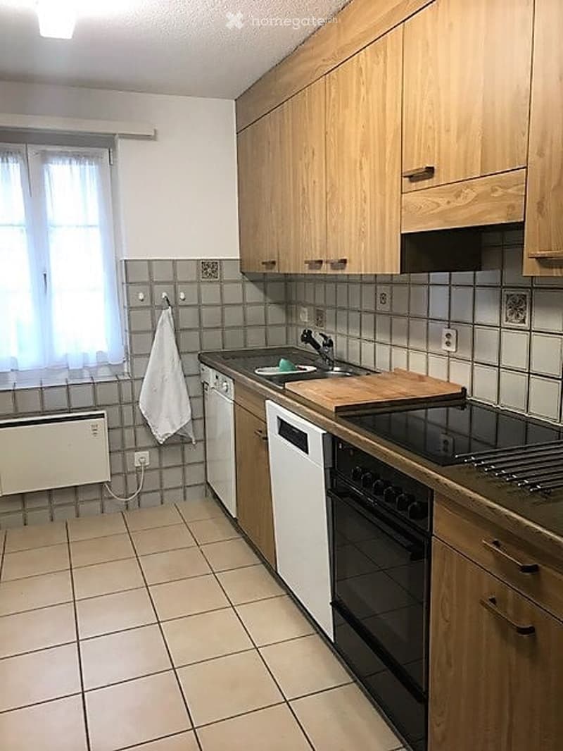 Modern kitchen with light wooden cabinets, tiled backsplash, and stainless steel appliances including a sink, oven, and dishwasher. A window with white curtains provides natural light.