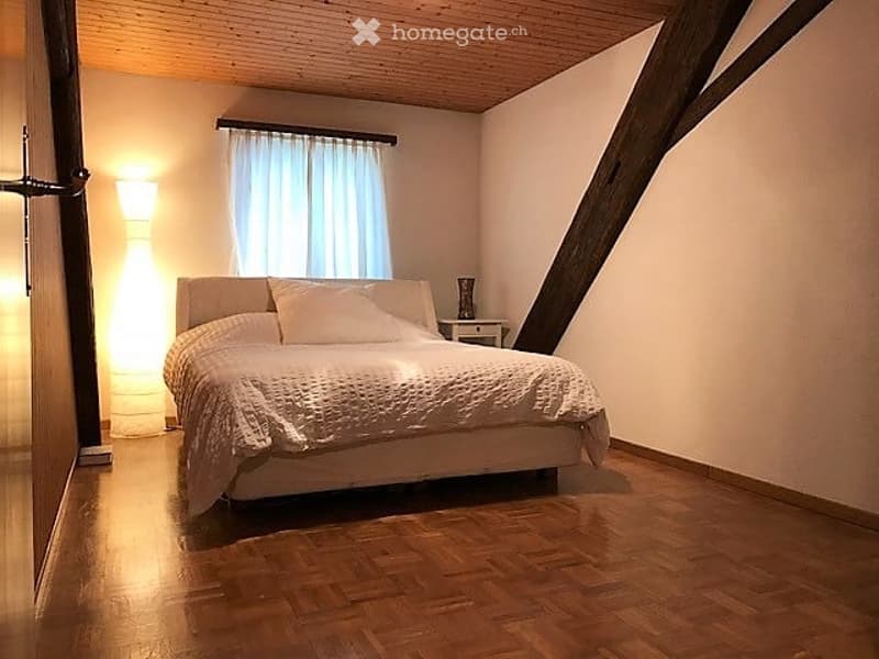 Cozy bedroom with wooden flooring, a large bed with white bedding, a nightstand with a lamp, and a window with white curtains.