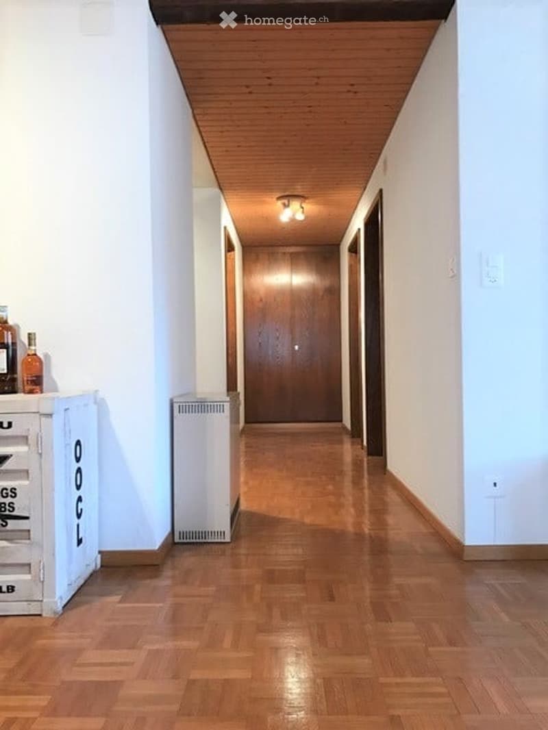 A narrow hallway with wooden flooring and white walls, featuring a wooden ceiling and recessed lighting. There are a few items on the left side, including a small cabinet and some bottles.