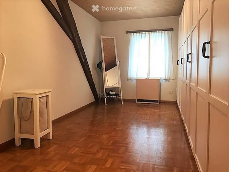 Spacious room with wooden flooring, large window with white curtain, wardrobe, mirror, and small stool.