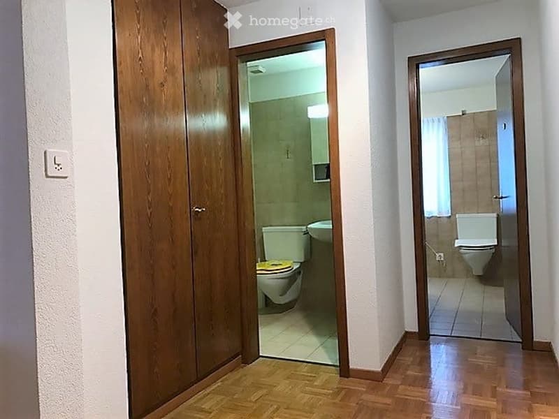 A narrow hallway with wooden flooring and white walls, featuring two open doors leading to bathrooms with visible fixtures.