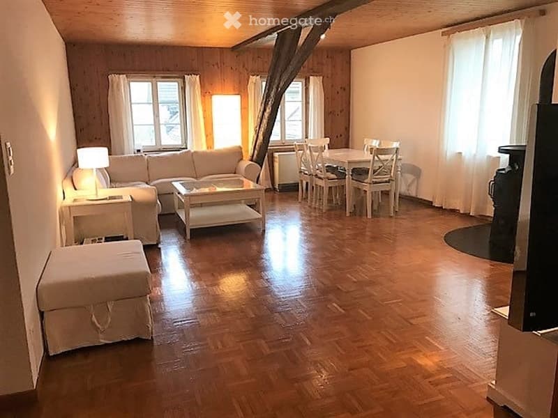Bright, open living and dining area with wooden floors, large windows, and exposed wooden beams. Features a white sectional sofa, coffee table, ottoman, side table with lamp, and a dining area with a table and chairs.