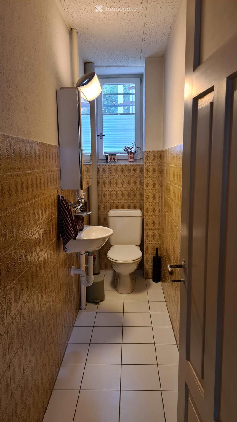 Small bathroom with beige tiles, white toilet, sink, and a window with blinds. A towel hangs on the wall, and a small plant sits on the windowsill.