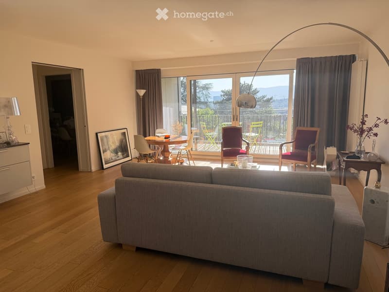 Modern living room with hardwood floors, large windows, grey sofa, coffee table, dining area with chairs and table, and decorative items.