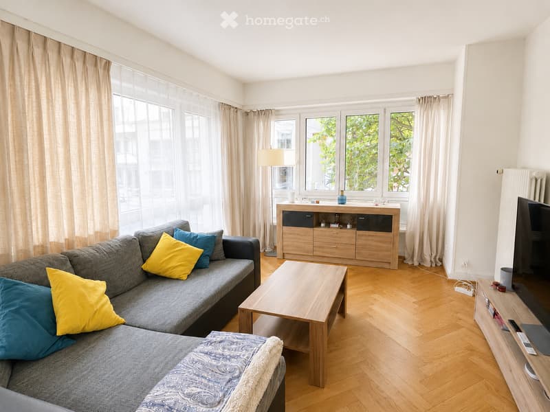 Modern living room with grey sofa, wooden coffee table, TV stand, floor lamp, and large windows with curtains.
