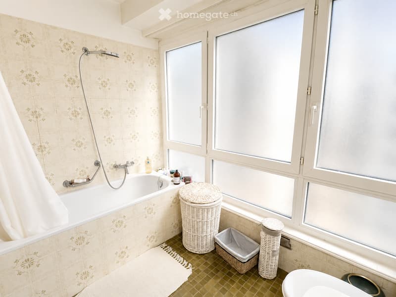 Bathroom with a bathtub, toilet, wicker baskets, and a trash bin. The walls are tiled with a floral pattern, and there is a large window providing natural light.