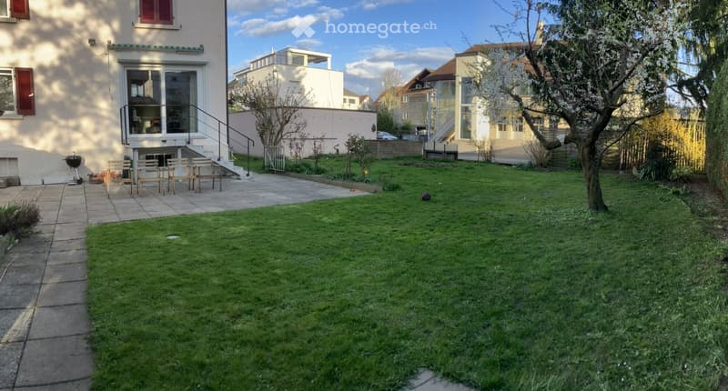 Well-maintained garden with a grassy lawn, patio area with chairs and a grill, and a modern house with a balcony in the background.