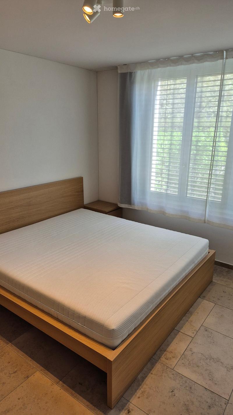 A bedroom with a wooden bed, white mattress, white walls, tiled floor, a nightstand, and a window with white curtains and blinds.