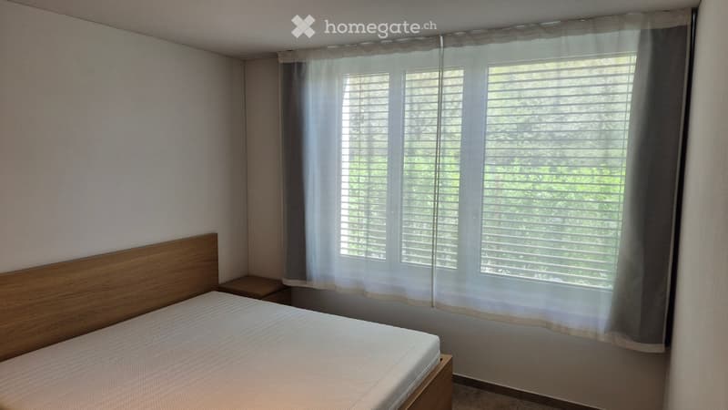 Bedroom with a wooden bed frame, a mattress, a nightstand, and a window with blinds and curtains.
