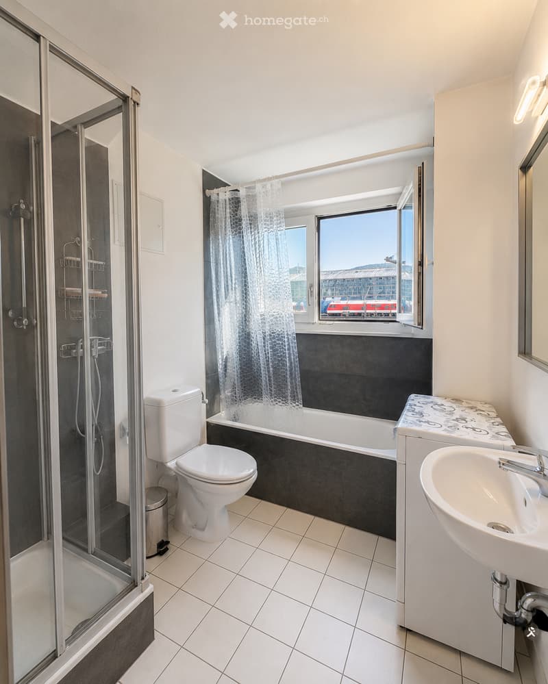 Modern bathroom with white tiles, a bathtub, toilet, sink, and a glass-enclosed shower. A large window offers a view of the city.