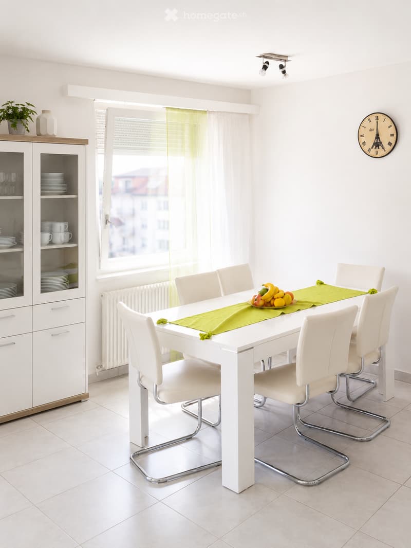 Bright dining room with white walls, a white dining table surrounded by six white chairs, a green table runner, and a fruit bowl. A white cabinet with glass doors holds dishes and cups, and a decorative clock is mounted on the wall. Natural light streams