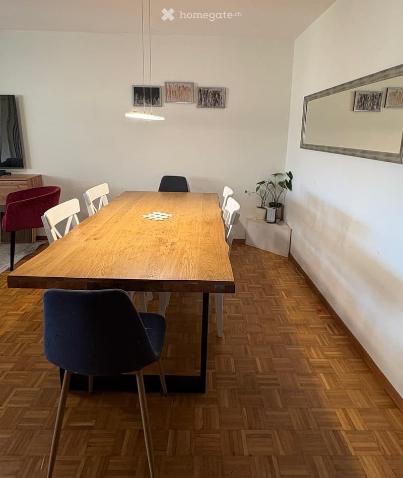 Dining room with a long wooden table surrounded by chairs, a potted plant on a small table, and artwork on the walls.